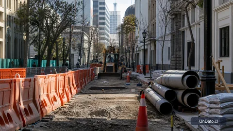 Calle urbana en reconstrucción con barreras de seguridad, materiales de obra y maquinaria al fondo.
