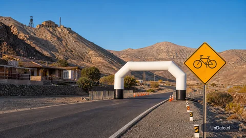 Camino rural en una localidad minera con señalización de una ruta ciclista, un arco de partida y cerros al fondo.