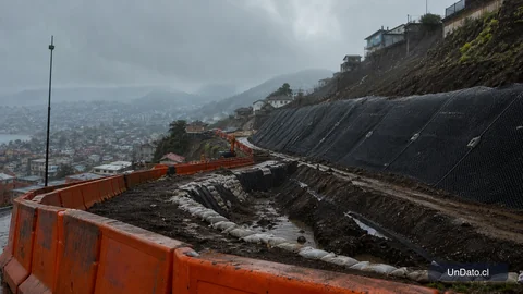 Obra vial con barreras de seguridad, sacos de arena, drenajes y una ladera bajo lluvia intensa.