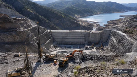 Obra de embalse en construcción con maquinaria pesada, terreno intervenido y marcas topográficas en un valle rural.
