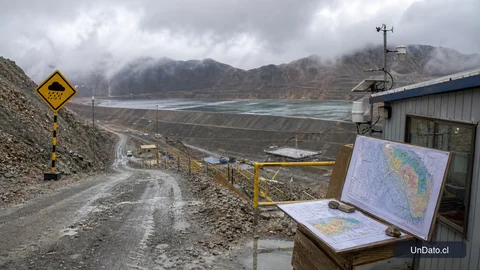 Ladera minera con camino de acceso mojado, señalética preventiva, un depósito de relaves y una caseta de monitoreo técnico.